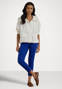 Svetla bunda s kapuco in dvema sprednjima žepoma, v kombinaciji z modrimi oprijetimi hlačami in rjavimi mokasini, z gladko teksturo.