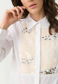White shirt featuring a front panel with reflective sequins and circular embellishments, contrasting with the smooth fabric of the collar.