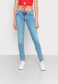 Vrouw in lichtblauwe skinny jeans, witte plateau sneakers en een rode cropped top die op de taille is geknoopt, staande tegen een egale achtergrond.