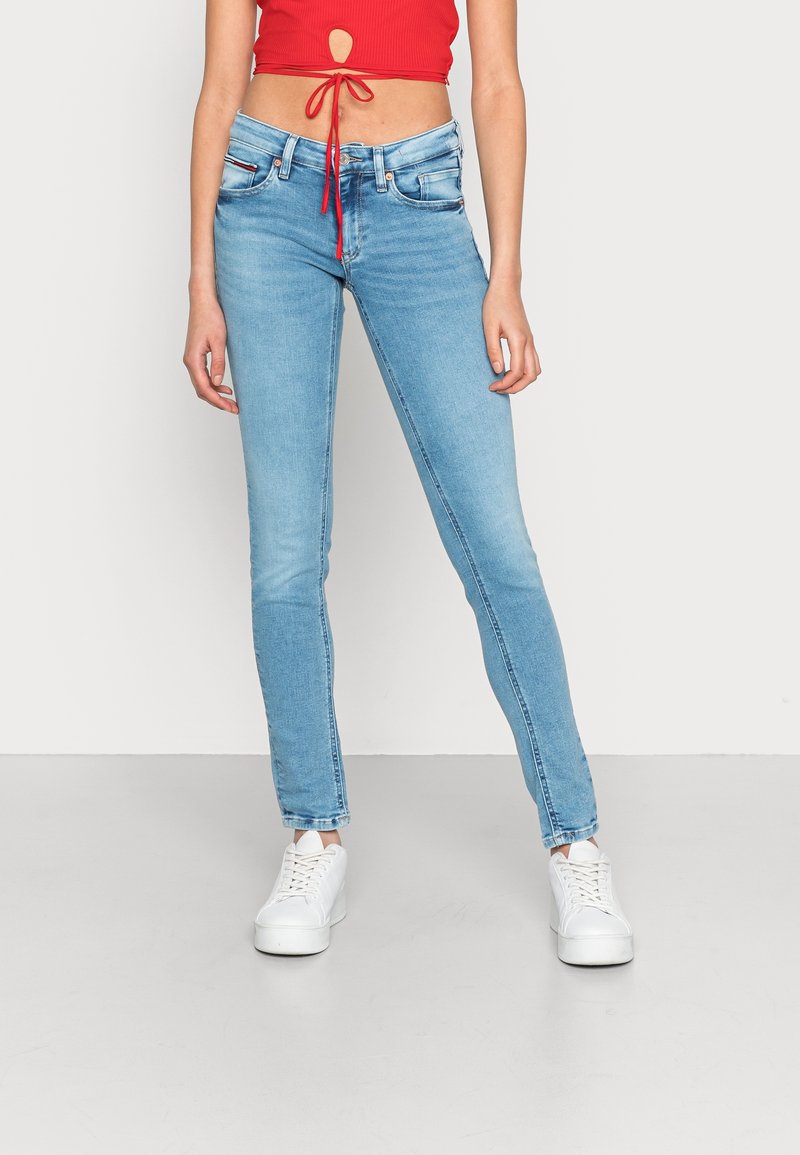 Vrouw in lichtblauwe skinny jeans, witte plateau sneakers en een rode cropped top die op de taille is geknoopt, staande tegen een egale achtergrond.