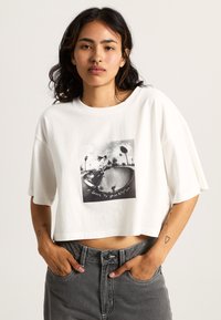 Witte cropped t-shirt van katoen, met een zwarte grafische print met de tekst "Skate to Your Rhythm" en palmbomen op een skateboard scène.