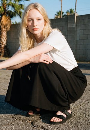 Jeune femme aux cheveux blonds accroupie en plein air, portant un t-shirt blanc, une jupe noire et des sandales noires à boucle près d'un mur en béton.