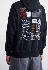 Svart huvtröja med grafiska tryck i vitt och orange. Designen inkluderar planeter, en astronaut och textfragment. Mjuk tygtextur.