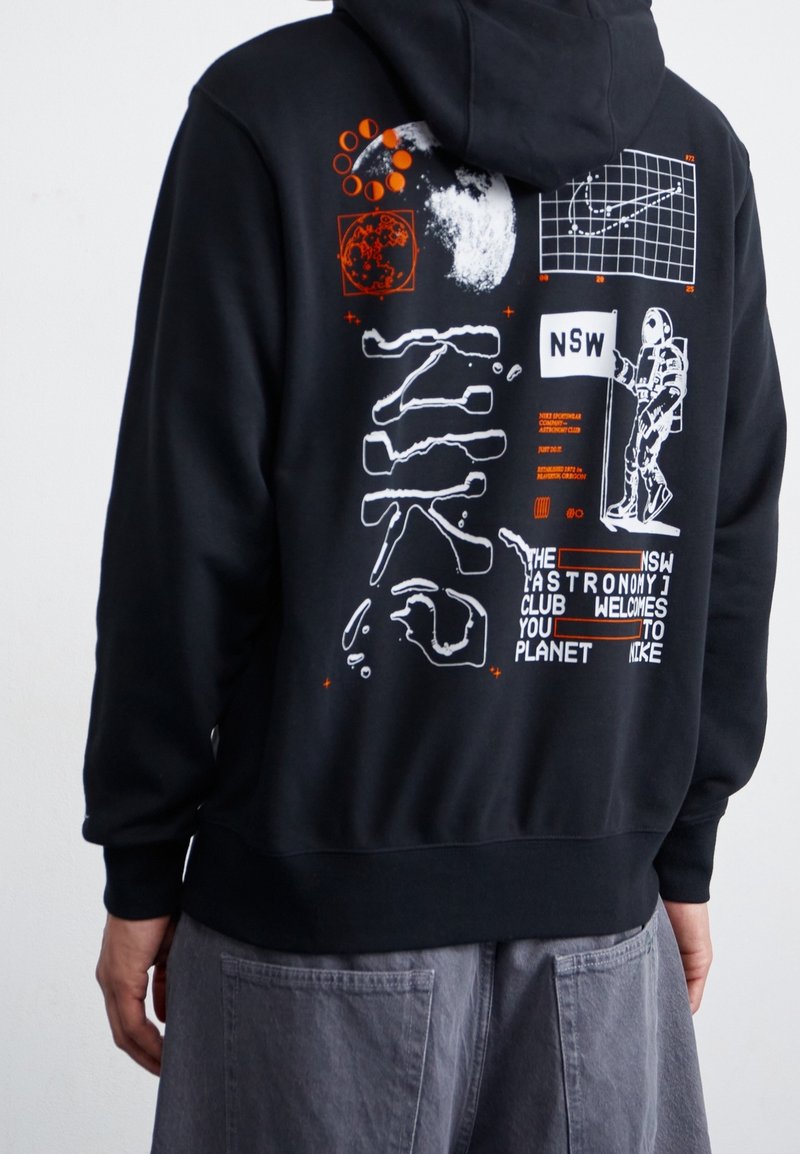 Svart huvtröja med grafiska tryck i vitt och orange. Designen inkluderar planeter, en astronaut och textfragment. Mjuk tygtextur.