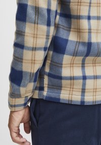 Fleece shirt in beige met een blauw en bruin geruit patroon, met een ronde zoom en een zijlabel. Materiaal lijkt zacht en gestructureerd.