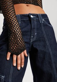 Svart nätad långärmad topp med lös väv, parat med mörkblå denimjeans som har kontrasterande vit söm och en rak benprofil.