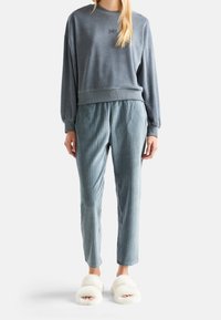 Person klädd i grå långärmad sweatshirt, ribbade blågrå byxor och vita fluffiga tofflor med öppen tå mot en neutral bakgrund.