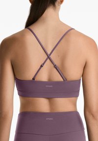 Soutien-gorge de sport violet avec des bretelles croisées réglables et une bande avec logo. Texture lisse, conçu pour le confort et le maintien durant les activités.