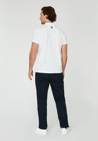 Weißes Poloshirt mit einem schwarzen Logo am Kragen, kombiniert mit dunkelblauen Hosen und weißen Sneakers. Das Shirt hat einen klassischen Kragen und kurze Ärmel.