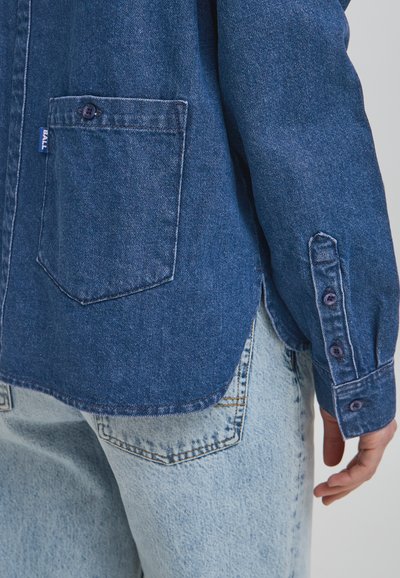 Jeansskjorta i mörkblått med en bröstficka, långa ärmar och knappmanschetter. Har en rundad nederkant och synliga sömdetaljer.