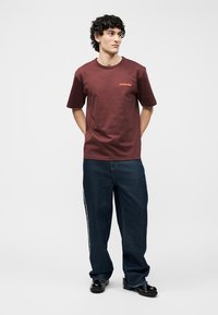 Νέος άνδρας με κατσαρά μαλλιά που φοράει μπορντό μπλουζάκι και χαλαρά σκούρα τζιν, στέκεται με τα χέρια πίσω από την πλάτη, κοιτάζοντας προς τα πλάγια.