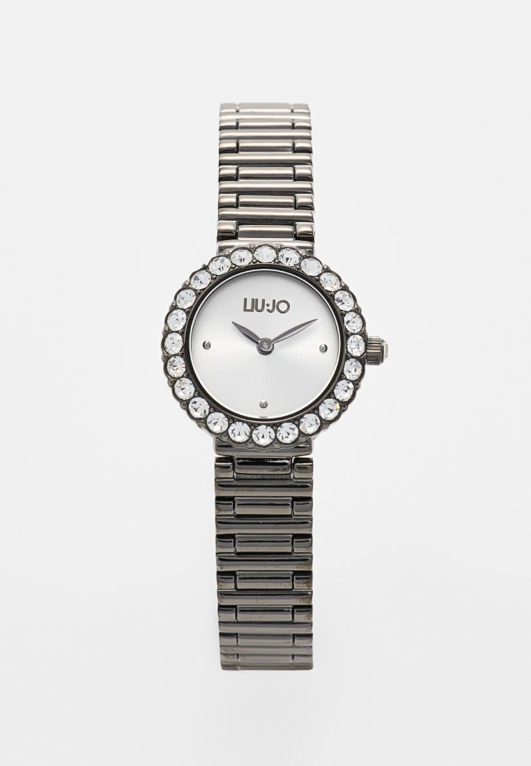 Silberne Uhr mit einem strukturierten Armband, rundem Ziffernblatt und Kristallakzenten um das Ziffernblatt. Sie hat einen weißen Hintergrund und schwarze Zeiger.