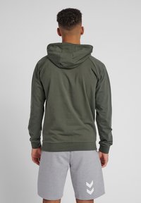 Olivgrön huvtröja med långa ärmar, ribbade muddar och en känguruficka. Bärs med ljusgrå shorts med vita sicksackmönster.