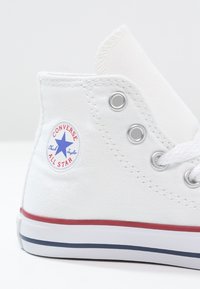 Bílé plátěné tenisky vysoké s stříbrnými očky, červeným a modrým pruhovaným podrážkou, s kulatým logem "Converse All Star" na boku.