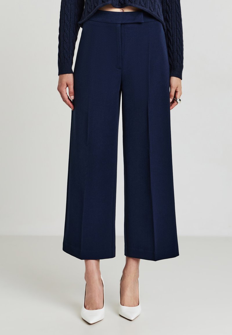 Marinefarbige Culottes mit glatter Textur, hoher Taille und figurbetonter Silhouette. Kombiniert mit weißen spitz zulaufenden Schuhen.