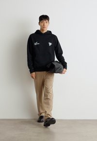 Haut à capuche noir avec impression sur le devant, pantalon beige et chaussures noires. Le modèle porte un sac noir texturé. Fond simple.
