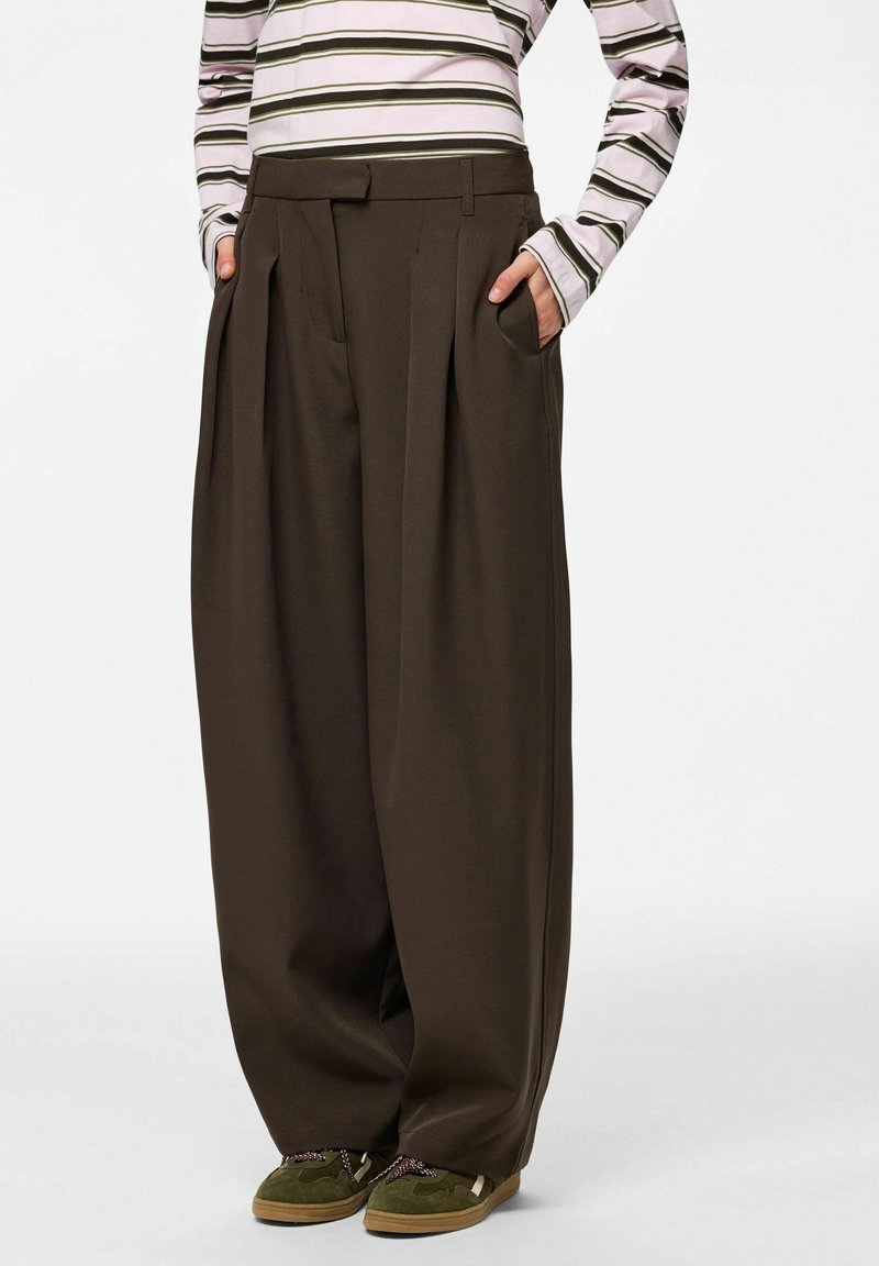 Personne portant un pantalon large marron foncé, un haut rayé à manches longues et des baskets vertes, les mains dans les poches.