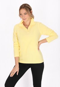 Gul fleece trøje med kvart lynlås, lange ærmer og krave. Glat tekstur, tætsiddende design i kombination med sorte leggings.