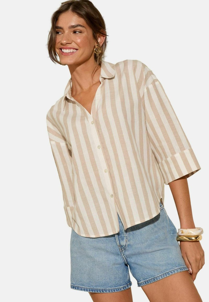 Camicia a maniche corte a righe in beige e crema, con una vestibilità comoda e un orlo leggermente curvo. Abbinata a pantaloni shorts di denim.