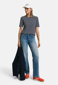Stribet marineblå bomulds-t-shirt, blå jeans, røde sneakers og en marineblå jakke. En kasket med hvid tekst og logo fuldender outfittet.