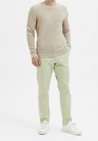 Beiger Strickpullover mit Textur und langen Ärmeln, kombiniert mit hellgrünen, gerade geschnittenen Hosen und weißen Sneakers. Lässiges Outfit, minimalistische Details.