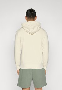 Hoodie beige clair avec capuche à cordon, manches côtelées et ourlet. Porté avec un short vert. Texture lisse, design décontracté.