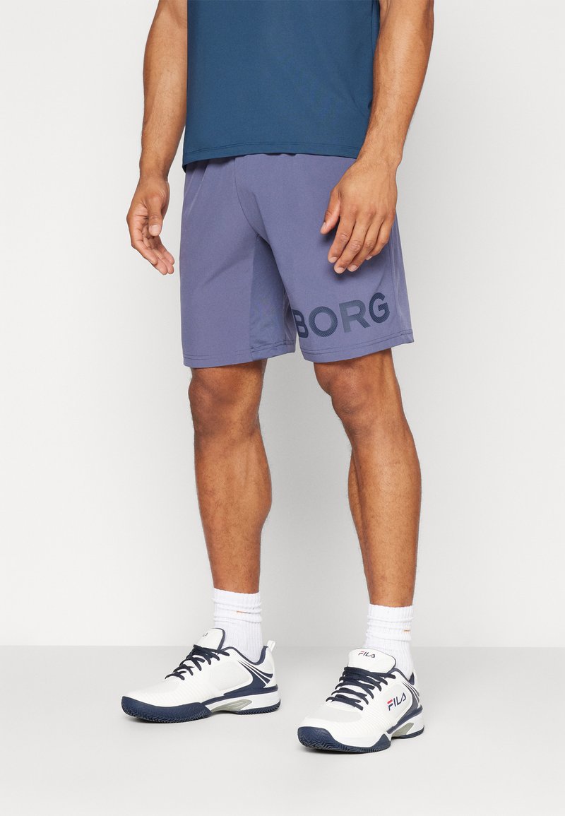 Pánske fialové športové šortky s veľkým čiernym logom "BORG", v kombinácii s bielymi a námornícky modrými športovými topánkami a bielymi členkovými ponožkami.