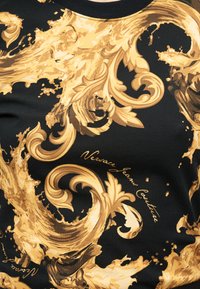 Μαύρο ύφασμα με περίτεχνες χρυσές βαρoκ σπείρες και το κείμενο «Versace Jeans Couture» σε καλλιγραφική γραφή ενσωματωμένο στο σχέδιο.