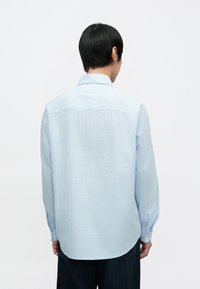 Jasnoniebieska koszula na guziki z fakturowanej tkaniny, z długimi rękawami, zaokrąglonym dołem i subtelnymi paskami, widoczna od tyłu.
