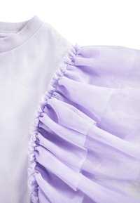 Lilafarget topp med en myk bomullsbody og en tulle-frynseerm. Frynsen har en rynket detalj langs kanten, noe som gir tekstur.