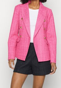 Světle růžový dvouřadý blazer vyrobený z texturované tkaniny se zlatými knoflíky, kombinovaný s tmavými tkanými šortkami.