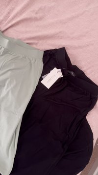 To par med bukser: en lys grå og en mørk sort, begge med elastisk linning og merkelapper festet, som hviler på en rosa teksturert overflate.