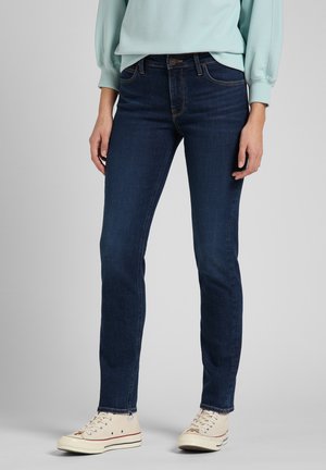 Person iført mørkeblå slim-fit jeans, lyseblå sweatshirt og off-white høje sneakers står mod en ensfarvet grå baggrund.