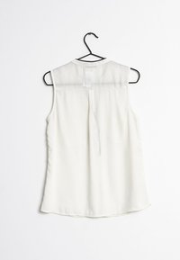 White sleeveless blouse with a sheer yoke and a round neckline. Features a back pleat and a curved hem. Hangs on a black metal hanger.
