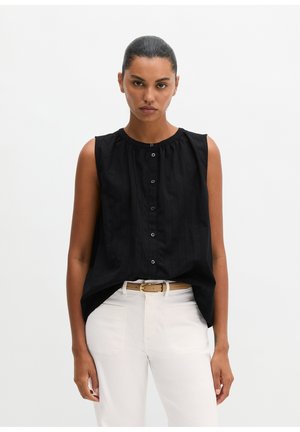 Frau mit zurückgebundenem Haar, die ein ärmelloses schwarzes, geknöpftes Oberteil und weiße Hosen mit beigem Gürtel trägt, steht vor einem einfarbigen Hintergrund.