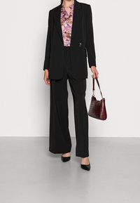 Blazer nero su misura con pantaloni larghi coordinati, indossato sopra una blusa floreale. Tiene una borsa bordeaux texturizzata e indossa tacchi neri.