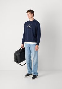 Sweatshirt azul-marinho com o logo "Calvin Klein", jeans Azul claro, ténis pretos e uma mala de desporto preta. Tecido suave, corte descontraído.
