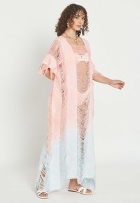 Mujer posando con un vestido largo con flecos degradados en tonos rosa y blanco, llevando sandalias blancas y un collar dorado sobre un fondo liso.