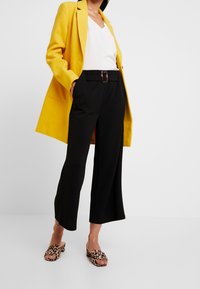 Blazer jaune, blouse blanche et pantalons larges noirs. Sandales à enfiler à imprimé léopard. Design ajusté avec une taille ceinturée.