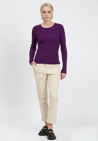 Lila gerippter Pullover mit langen Ärmeln und rundem Ausschnitt, kombiniert mit cremefarbigen maßgeschneiderten Hosen und schwarzen Slippern mit goldenen Akzenten.