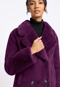 Fioletowy płaszcz z sztucznego futra o podwójnym zapięciu, z dużymi klapami i czarnymi guzikami. Długie rękawy i miękka, teksturowana powierzchnia.