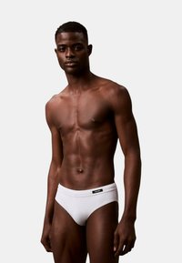 Slip de bain blancs avec une texture lisse et une taille élastique, arborant une petite étiquette de logo noire. Le modèle a une silhouette athlétique.