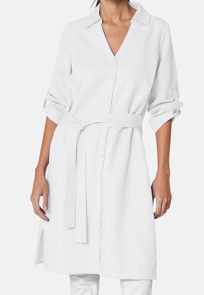 Model wearing a white, button-up shirt dress with rolled sleeves and a tied belt at the waist, showcasing a sleek, minimalist design.