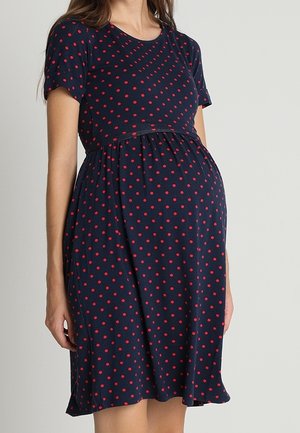 Pregnant woman wearing a navy blue dress with red polka dots, short sleeves, and a gathered waist, standing against a plain background.