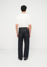 Άνδρας στέκεται με την πλάτη σε έναν λευκό τοίχο, φοράει λευκό T-shirt, σκούρα μπλε τζιν και μαύρα αθλητικά παπούτσια με λευκές ρίγες.