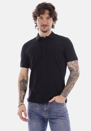 Uomo con capelli ricci, indossa una polo nera e jeans blu, mostra tatuaggi su entrambi gli avambracci, in piedi con una mano in tasca.