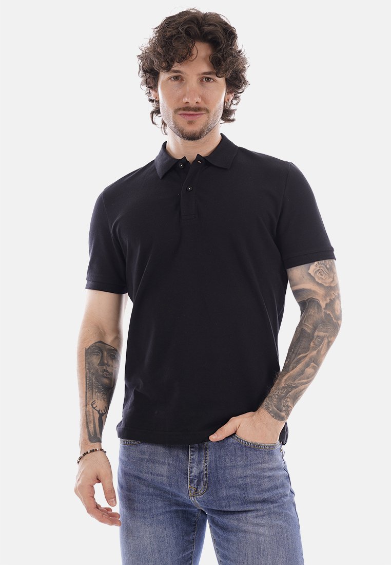 Hombre con cabello rizado, vestido con una camisa tipo polo negra y jeans azules, mostrando tatuajes en ambos antebrazos, de pie con una mano en el bolsillo.