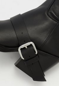 Black leather ankle boot with a chunky heel, featuring a broad strap secured by a silver buckle. Textured surface and angular design details.