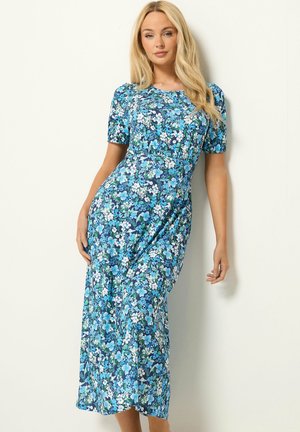 Femme aux longs cheveux blonds portant une robe midi bleue à imprimé fleuri et manches courtes, debout devant un fond uni clair.