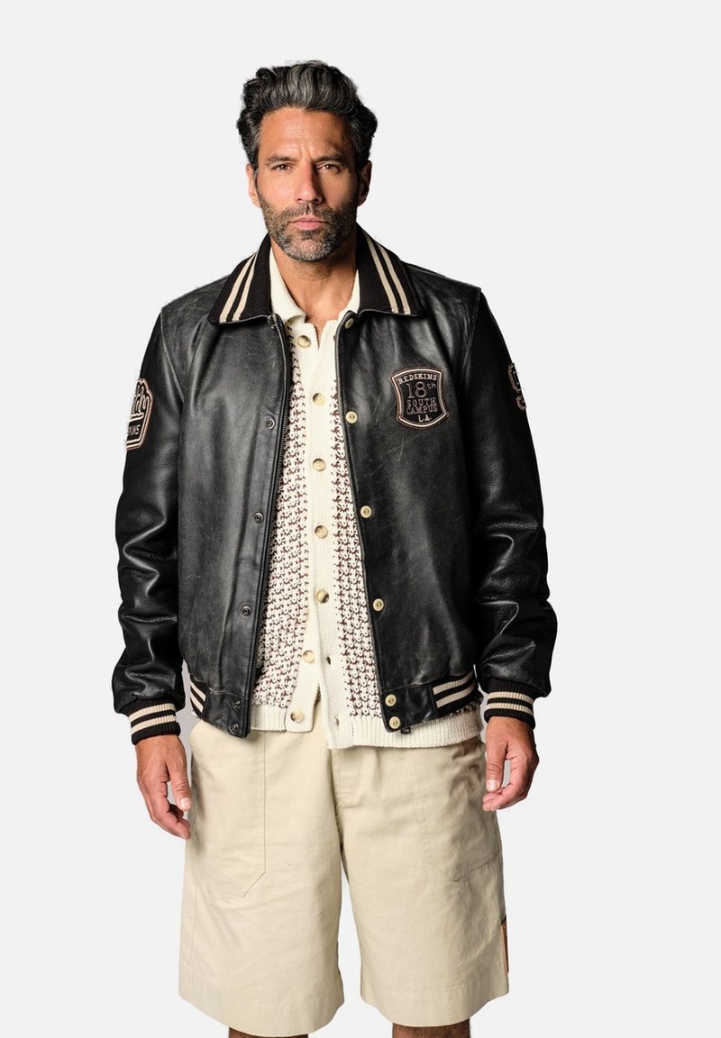 Veste universitaire en cuir noir avec col et poignets rayés, ornée de patchs, portée sur un pull en maille crème et un short beige.